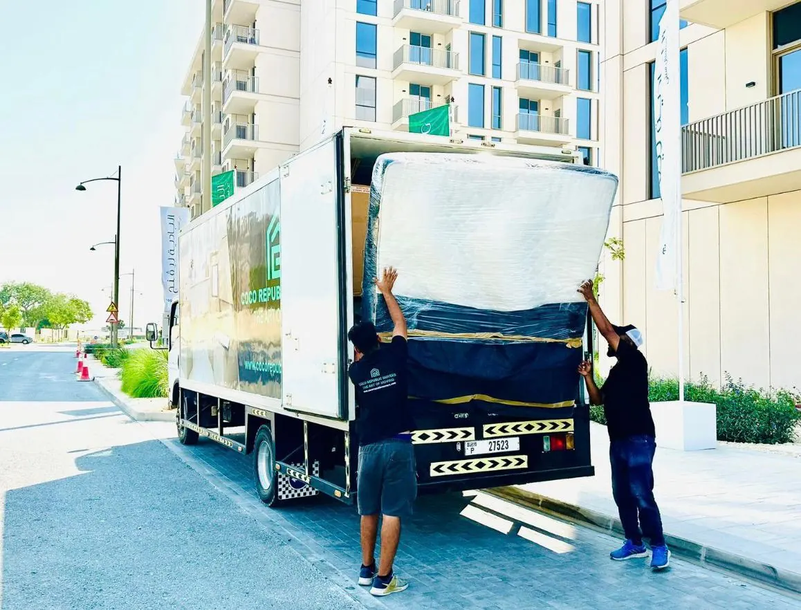 Two movers loading a wrapped mattress into a moving truck outside a Dubai apartment building
